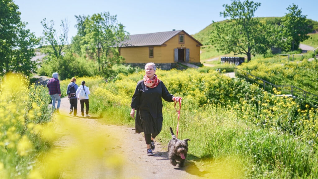Nainen ulkoiluttaa koiraa hiekkapolulla. Taustalla näkyy kolme ihmistä, keltainen rakennus, vihreää ja keltaista kasvillisuutta sekä sininen taivas.