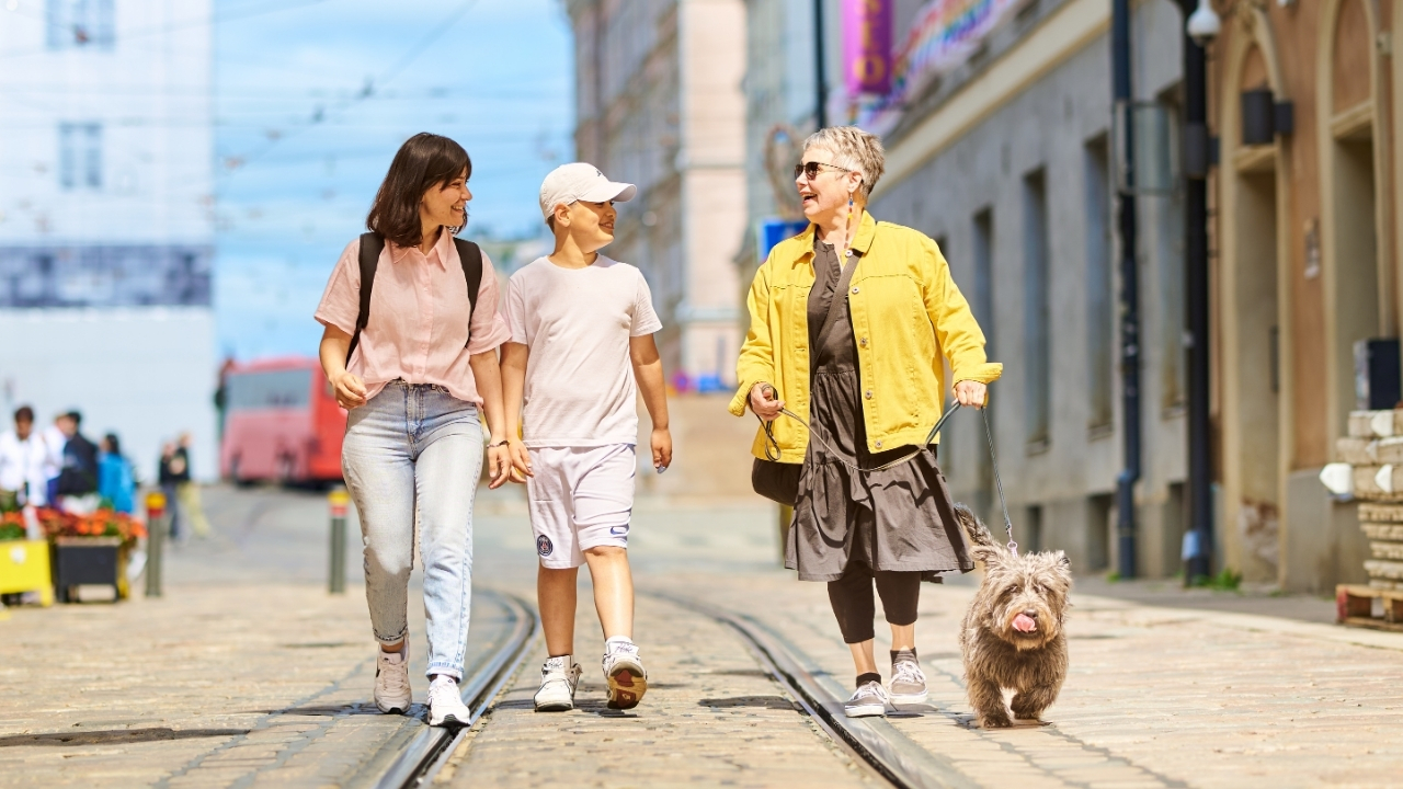 Nainen, poika, nainen ja koira kävelevät aurinkoisella kadulla. Taustalla näkyy rakennuksia ja sinistä taivasta.