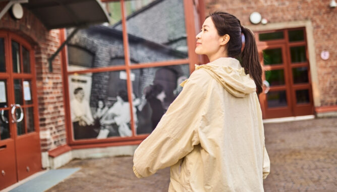 Nainen seisoo ja katse on suunnattu sivulle kauas. Taustalla punatiilinen rakennus ja ovia.