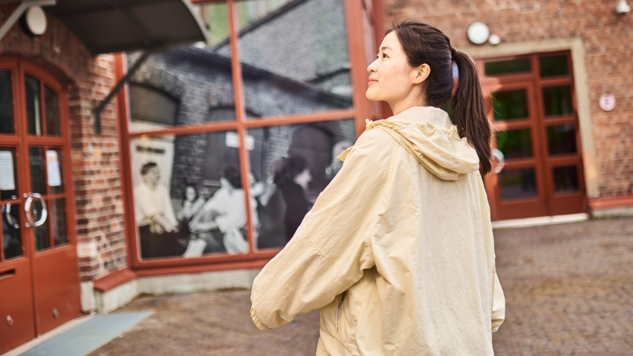 Nainen seisoo ja katse on suunnattu sivulle kauas. Taustalla punatiilinen rakennus ja ovia.
