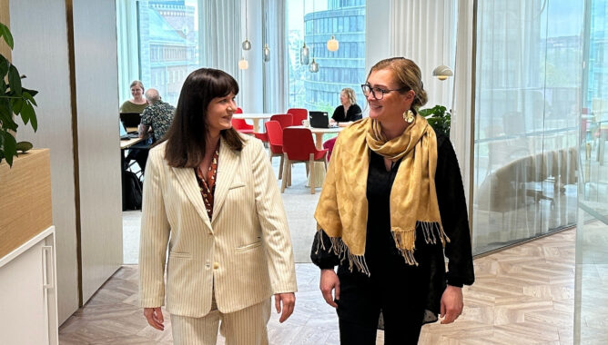 Kaksi naista kävelee toimistossa ja katsoo toisiaan. Taustalla näkyy ihmisiä pöytien ääressä.