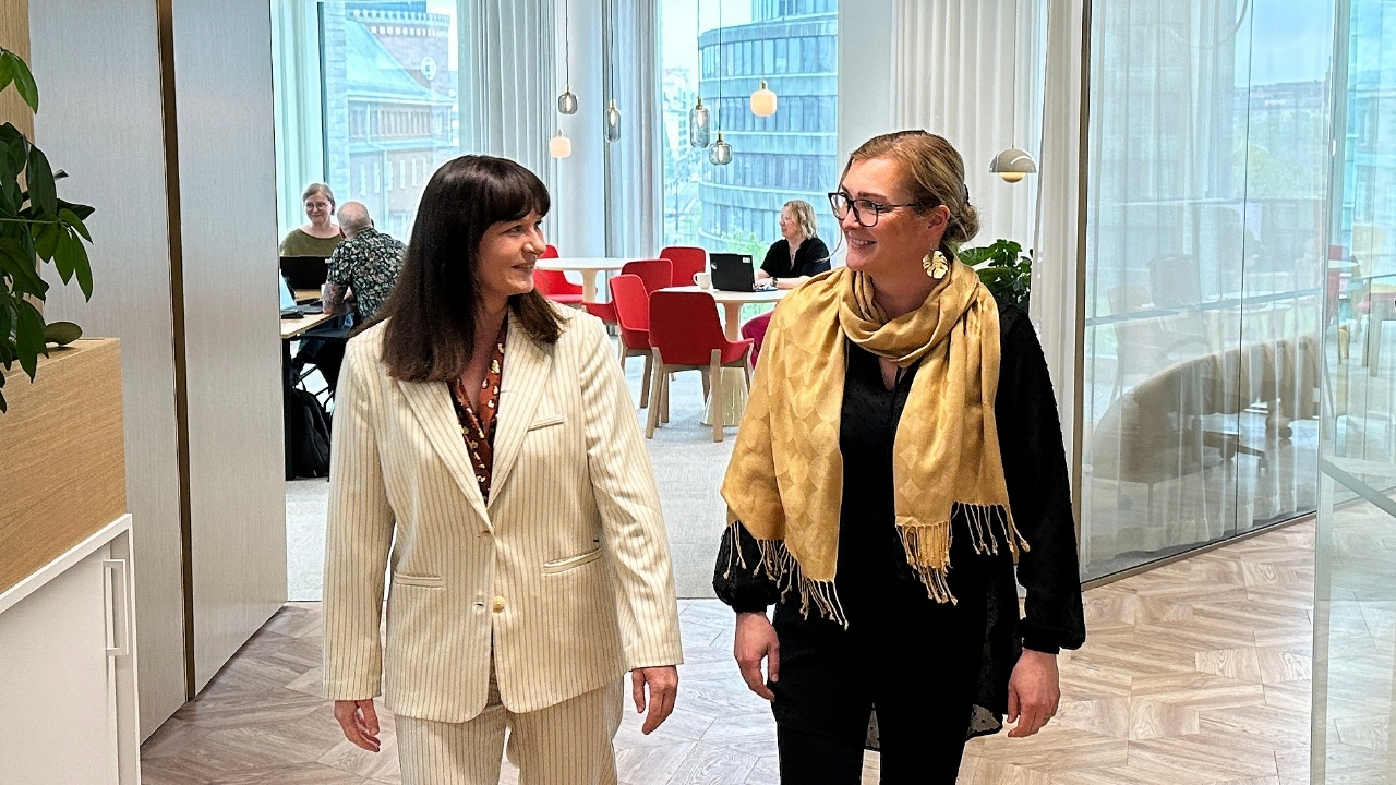 Kaksi naista kävelee toimistossa ja katsoo toisiaan. Taustalla näkyy ihmisiä pöytien ääressä.